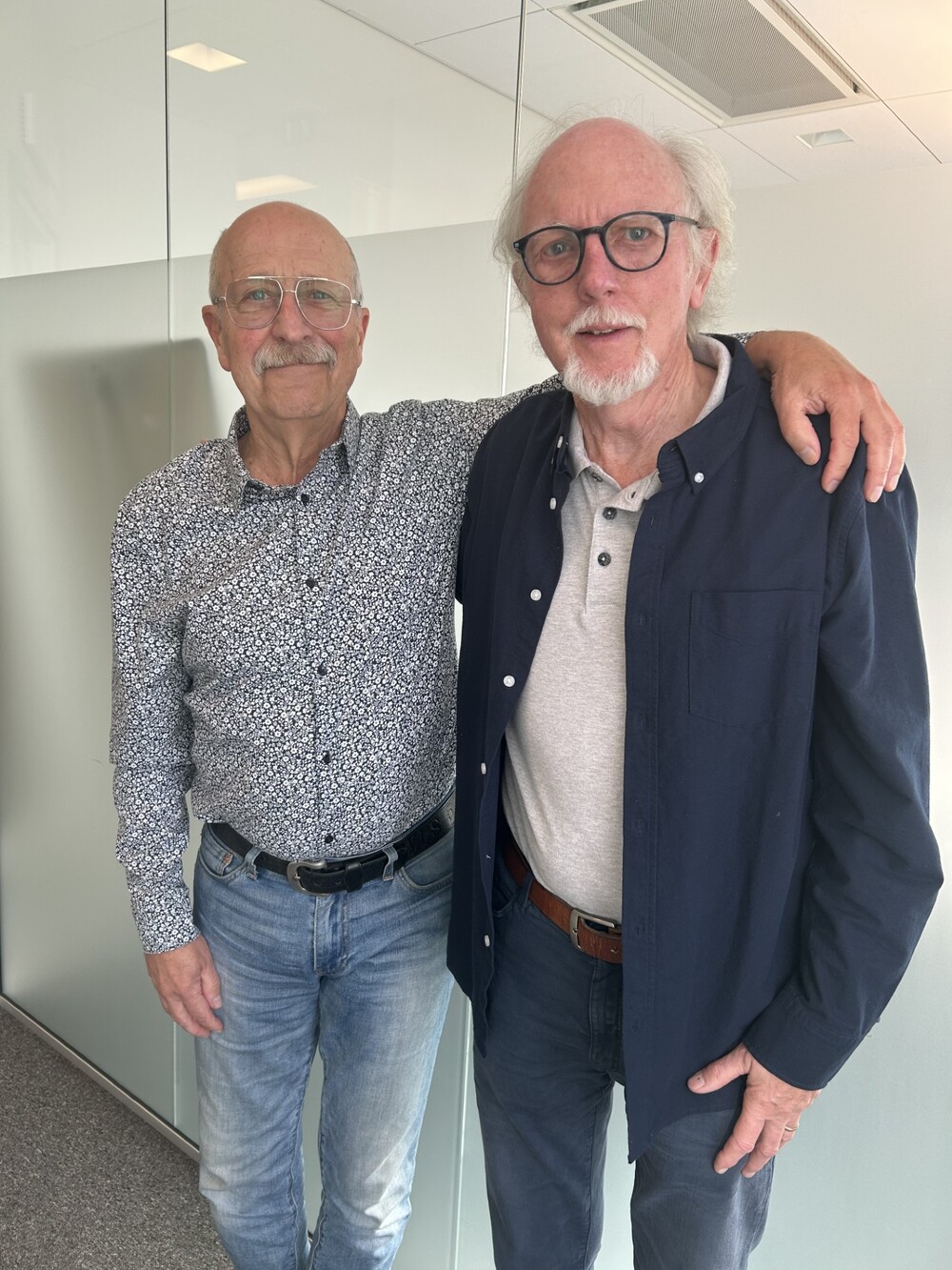 Två äldre män står bredvid varandra, den ena med armen runt den andra, i ett ljust kontorsrum.