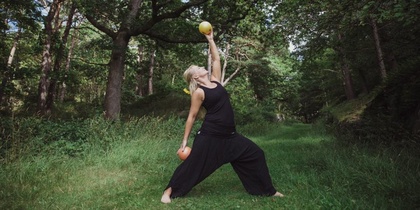 Person i svarta kläder gör yoga eller stretching med frukt i händerna i en grön skog.