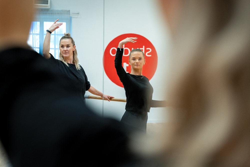 Två kvinnor övar dans framför en spegel i en dansstudio, båda med en arm upplyft.