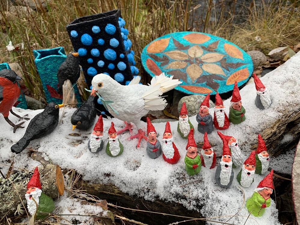 Flera små tomtefigurer och fåglar står på en snötäckt trädstam med färgglada dekorationer i bakgrunden.