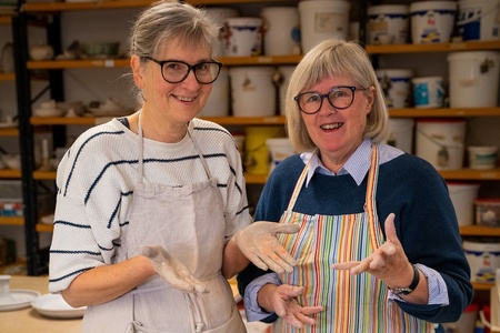 Två leende kvinnor med glasögon och förkläden står i en keramikverkstad med händerna täckta av lera.
