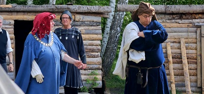 Tre personer i medeltida kläder står framför en timrad byggnad utomhus.