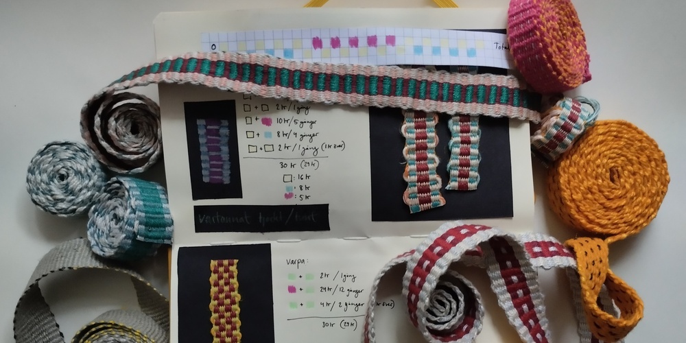 Flera vävda band i olika färger och mönster ligger upprullade på ett bord med en anteckningsbok med vävnoteringar.