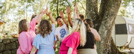 En grupp människor står i en ring utomhus och sträcker upp händerna mot mitten under ett träd.