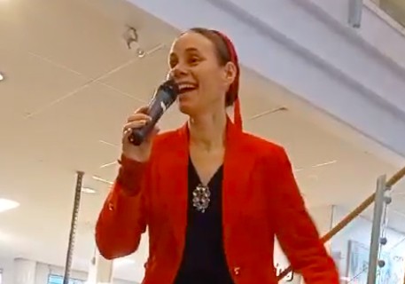 Person i röd kavaj håller mikrofon och talar inomhus, med modern inredning i bakgrunden.