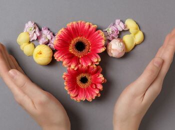Två händer omger blommor arrangerade som en livmoder på grå bakgrund.