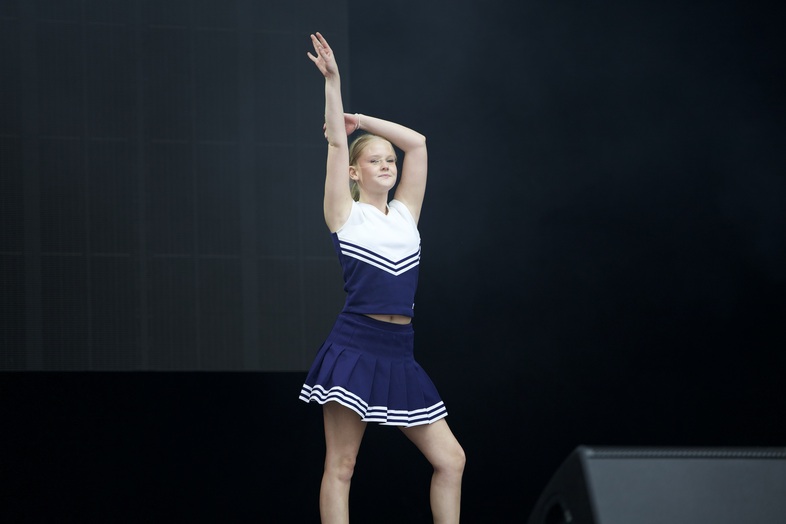 En ung kvinna i blå och vit cheerleaderdräkt står på scen med ena armen uppsträckt.