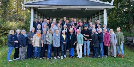 En stor grupp människor står utomhus framför en liten scen i en grönskande park.
