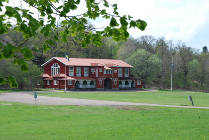 Röd trävilla med vita detaljer och svenska flaggor, omgiven av grön gräsmatta och träd.