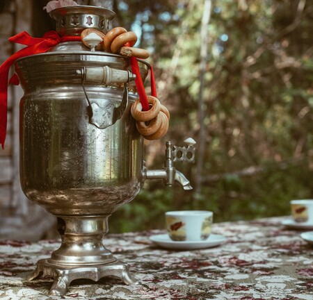 En samovar med röda band och kringlor står på ett bord utomhus, med två tekoppar bredvid.