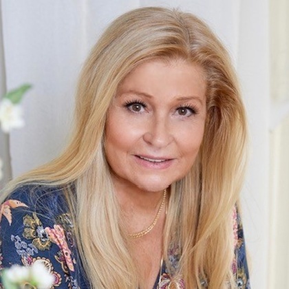 Kvinna med långt blont hår och blommig blus sitter inomhus och ler mot kameran.