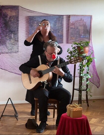 En person spelar gitarr sittande medan en annan står bakom och håller en blomsterkrans. Inomhusmiljö med växter och present.