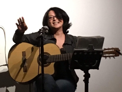 Kvinna sitter och spelar gitarr, sjunger i mikrofon och ler mot publiken.