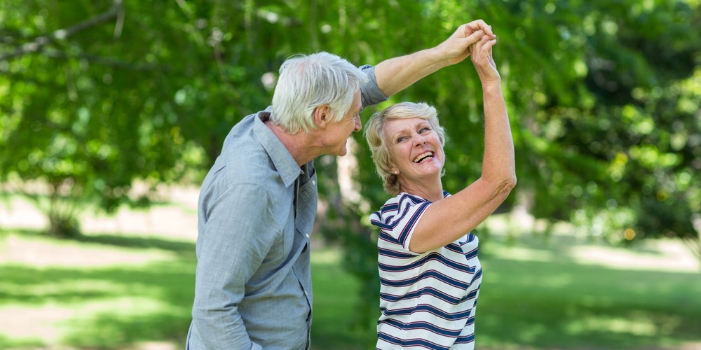 Ett äldre par dansar och skrattar tillsammans utomhus i en grönskande park.