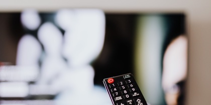En hand håller en fjärrkontroll riktad mot en tv med suddig bild i bakgrunden.