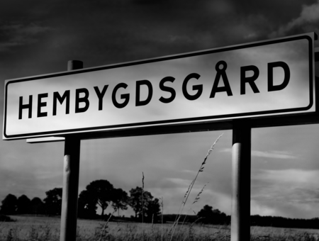 Vägskylt med texten 'HEMBYGDGÅRD' mot en landsbygd med träd och fält under en dramatisk himmel.