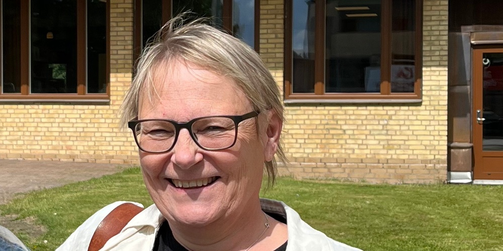 Två leende kvinnor står framför en gul tegelbyggnad med skylten 'BIBLIOTEK' under en blå himmel.