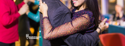 Två personer dansar nära varandra på en social tillställning, leende och festlig stämning.