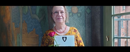 Kvinna med glasögon håller ett diplom och en röd ros, står framför en färgglad vägg och ett fönster.