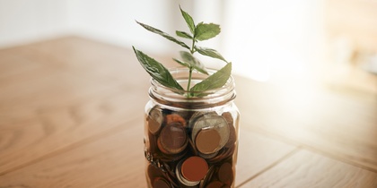 En glasburk fylld med mynt står på ett träbord, med en grön växt som växer ur burken.