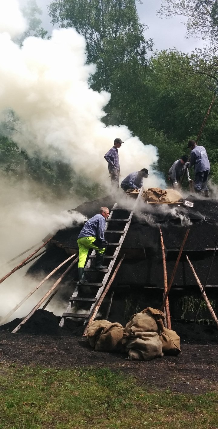 Flera personer arbetar på en rykande kolmila utomhus omgiven av träd och säckar.