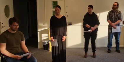 Fyra personer står och sitter i ett klassrum, tre står upp och en sitter ner, alla håller papper.