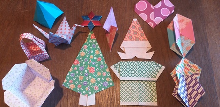 Flera färgglada origamifigurer, inklusive en julgran, hjärta, korg och lådor, ligger på ett träbord.