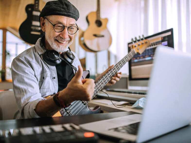 En person med grått skägg och bakvänd keps sitter och spelar elgitarr framför en laptop.