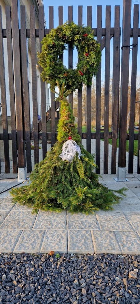 En dekoration av granris formad som en stor grön grinch med en krans som huvud, placerad utomhus framför ett staket.