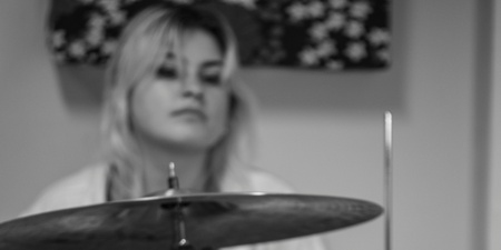 Svartvit bild av en kvinna som spelar trummor, med ett förstärkarhuvud i förgrunden.