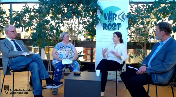 Fyra personer sitter i en paneldiskussion inomhus med gröna växter och en banner i bakgrunden.