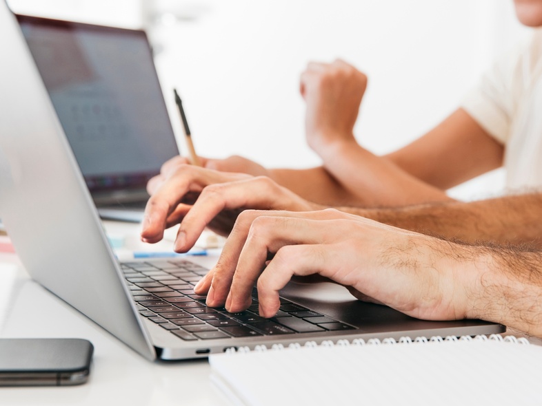 Två personer arbetar vid en bärbar dator med anteckningsblock och mobiltelefon på ett vitt skrivbord.