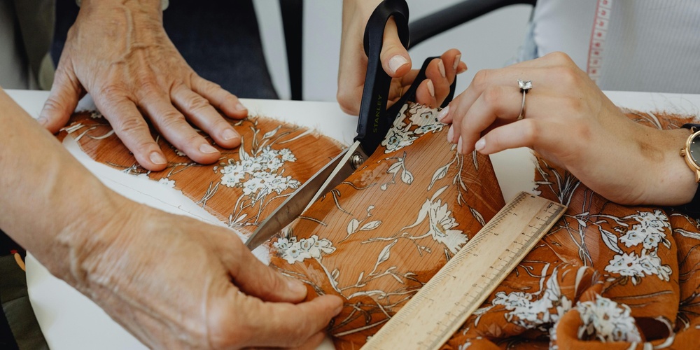 Två personer klipper ett blommigt tyg med sax och mäter med linjal på ett bord.