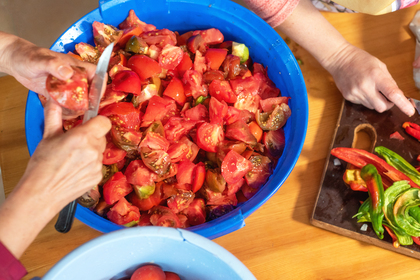 Två personer skär tomater och paprika vid ett bord, med en stor blå skål fylld med hackade tomater.