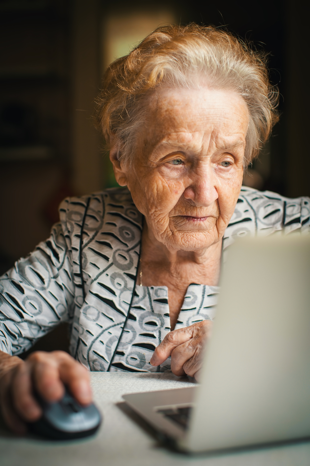 Äldre kvinna sitter vid ett bord och använder en bärbar dator med en datormus.