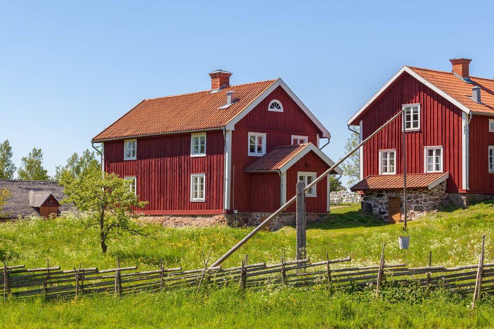 Två röda trähus med vita knutar på landet, omgivna av gräs, staket och blå himmel.