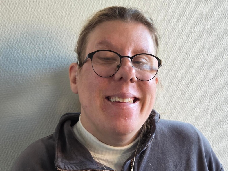 Person med suddat ansikte bär grå tröja och halsband, står mot ljus vägg.