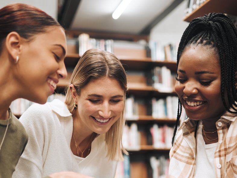 Tre unga kvinnor sitter på golvet i ett bibliotek och läser tillsammans ur en bok.