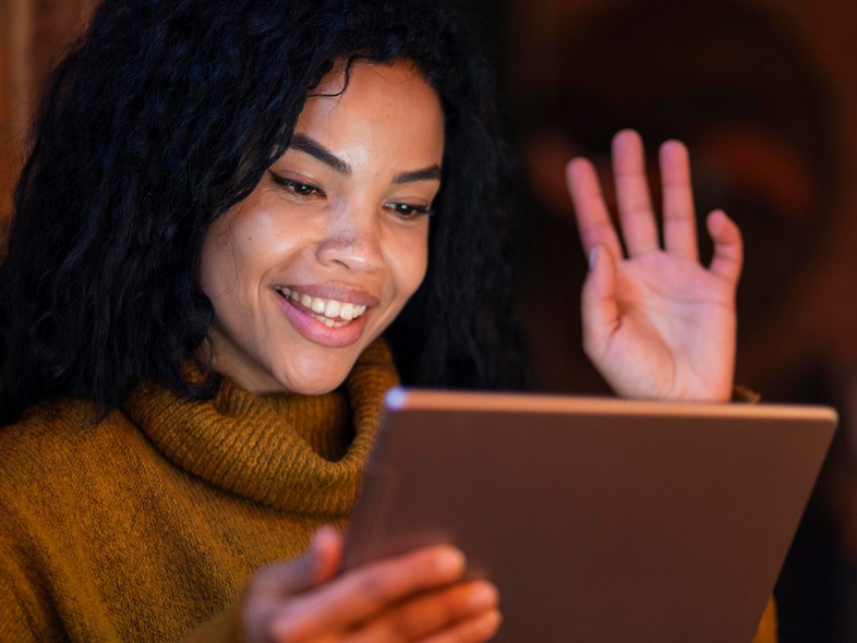 Kvinna med lockigt hår vinkar och ler mot en surfplatta i ett varmt upplyst rum.