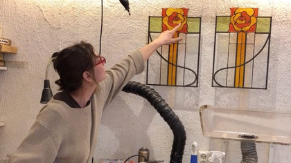 En kvinna pekar på ett färgglatt glasfönster med rosmotiv i en verkstadsmiljö.