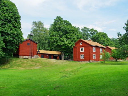Röda trähus omgivna av grönskande träd och gräsmatta under blå himmel.