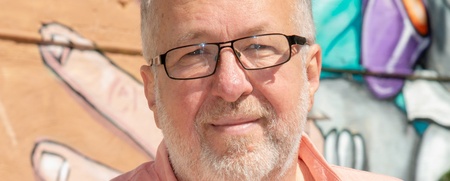 Äldre man med glasögon och skägg står framför en färgglad graffitivägg utomhus.