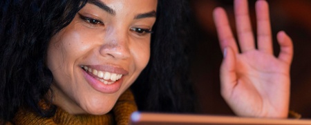 Kvinna med lockigt hår vinkar och ler mot en surfplatta i ett varmt upplyst rum.