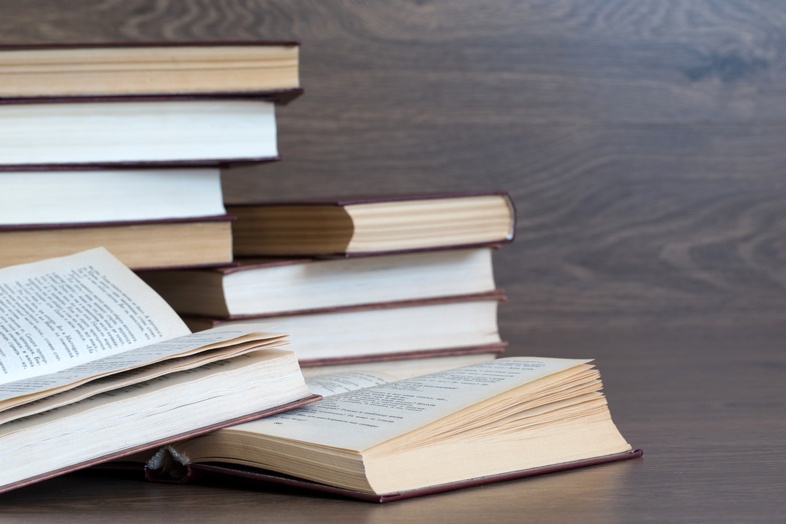 Flera böcker ligger och staplas på ett träbord, några är öppna och andra stängda.