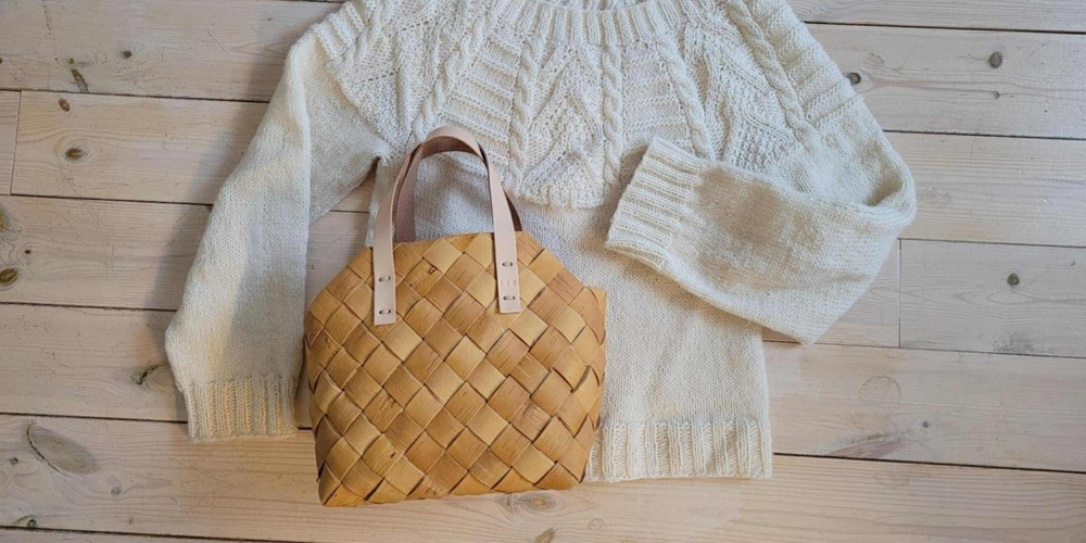 En vit stickad tröja och en flätad korgväska ligger på ett ljust trägolv.