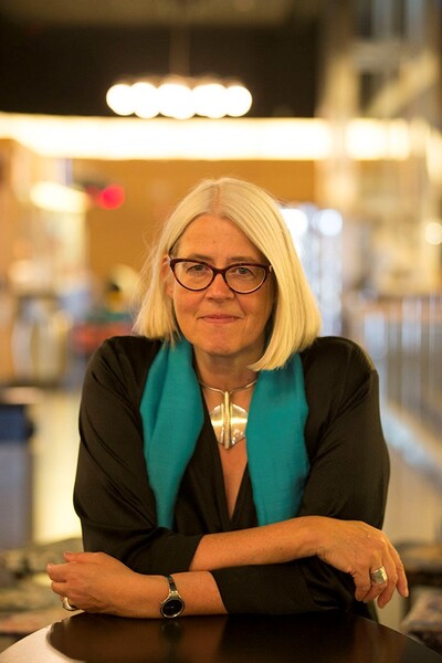 Person med blont hår och turkos sjal sitter vid ett bord, ansiktet är suddat.