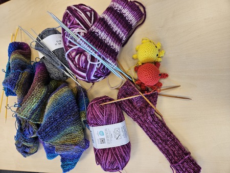 Flera stick- och virkprojekt med garn i olika färger, stickor och två små virkade figurer på ett bord.