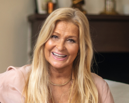 Kvinna med långt blont hår sitter i ett vardagsrum med mjuka färger och guldiga detaljer.