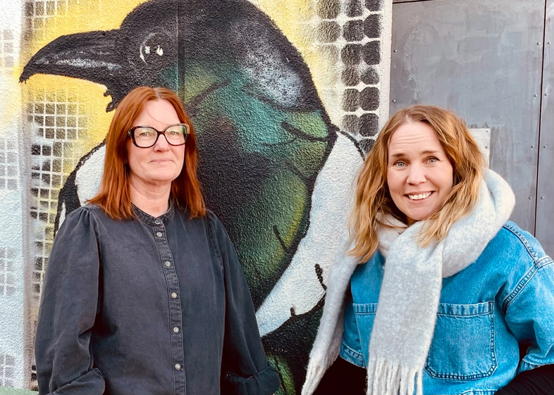 Två kvinnor står framför en väggmålning av en fågel, båda tittar mot kameran.