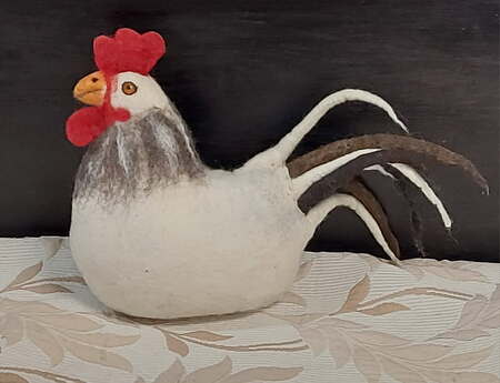 En filtad tupp i vitt, rött och brunt står på ett mönstrat tyg framför en mörk bakgrund.